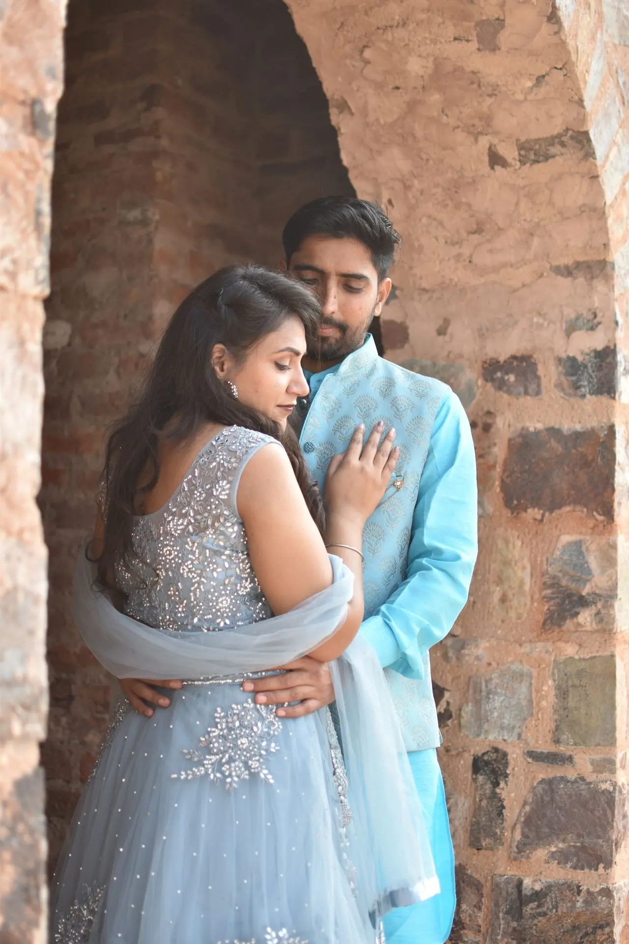 Couple wearing bright luxe matching light blue ethnic outfits leaning against a textured brick wall.