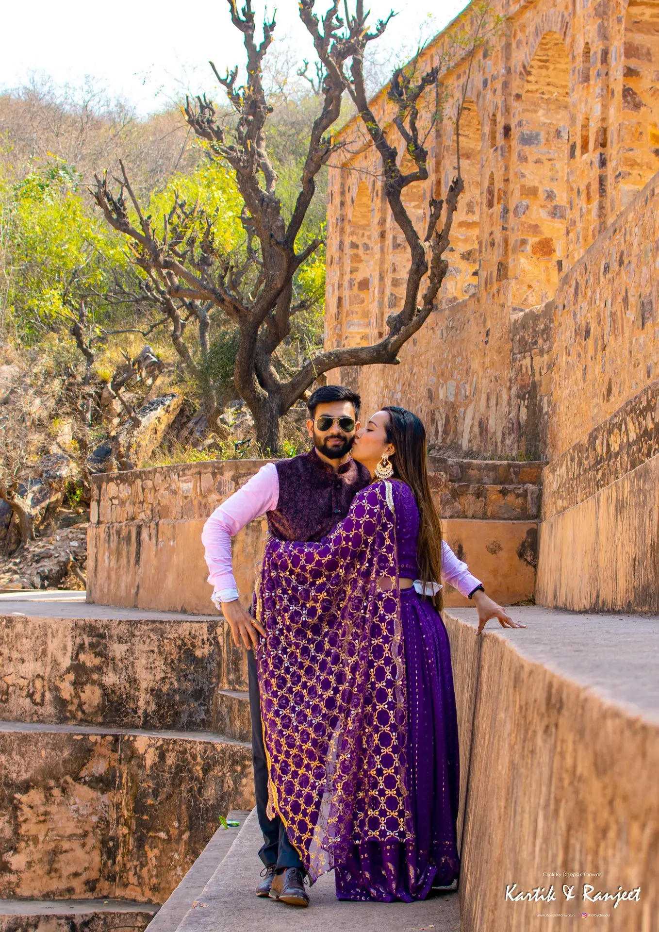 Wide shot of a couple in bright luxe turquoise and silver traditional wear sitting on a wall in a garden.