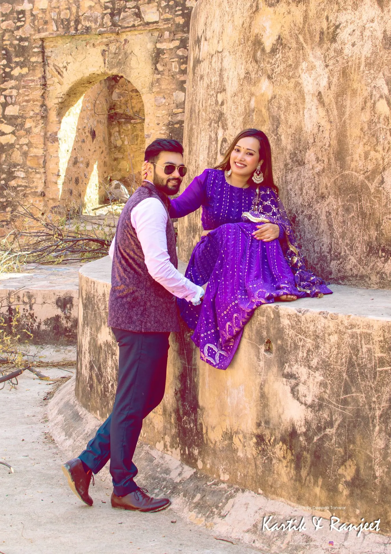 Couple posing in bright luxe purple traditional wear against a historic stone wall background.